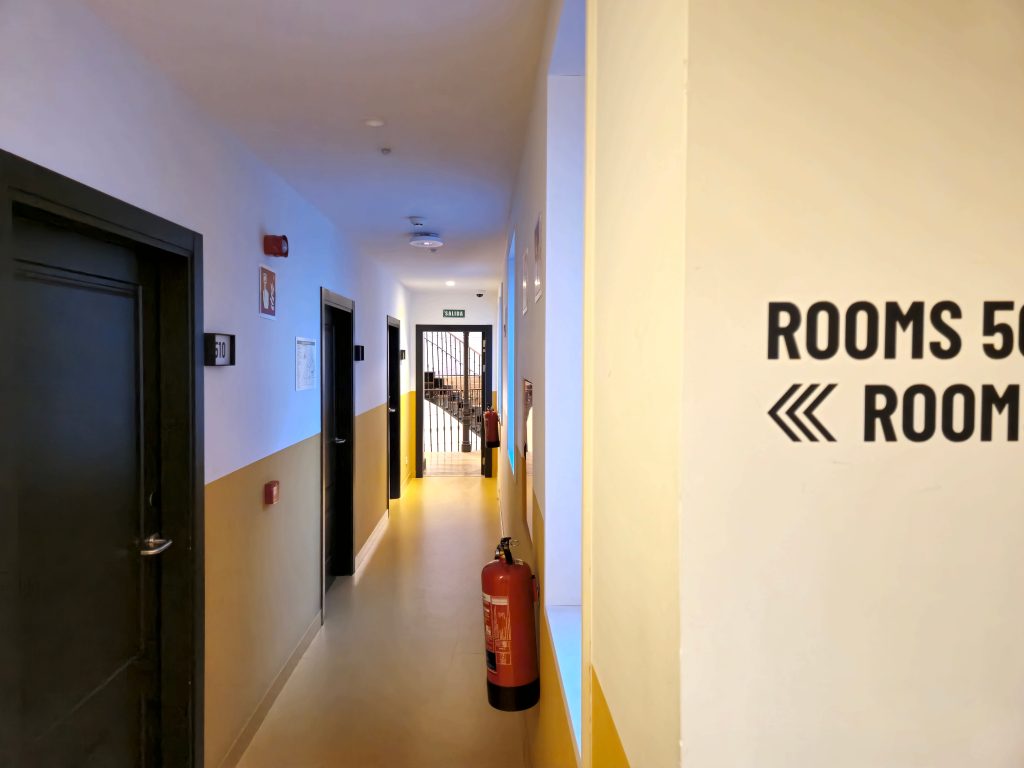 Long hotel corridor featuring sleek black doors lining the walls, a vibrant yellow floor guiding the way, and a fire extinguisher ensuring safety and security for guests